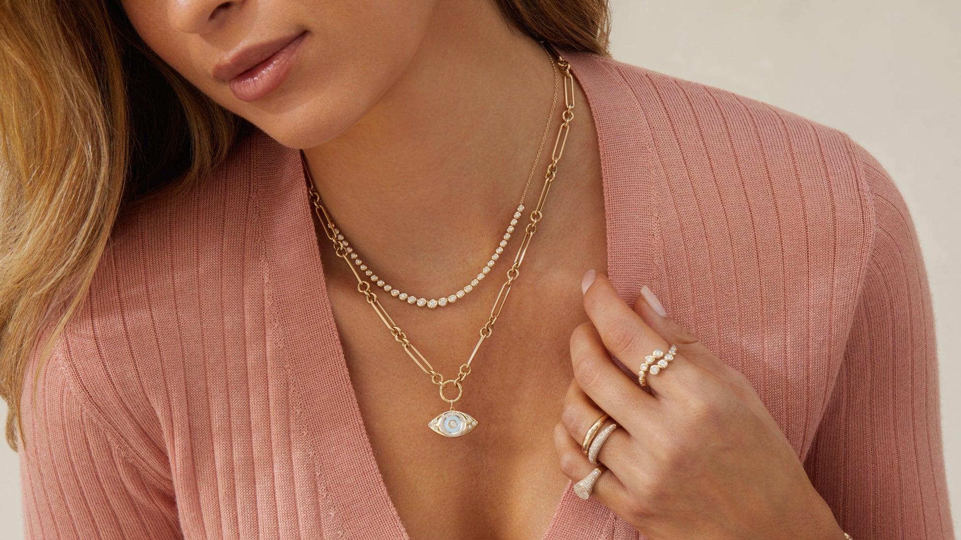 Woman with long blonde hair and a pink sweater wearing EF Collection layered necklaces and rings on multiple fingers
