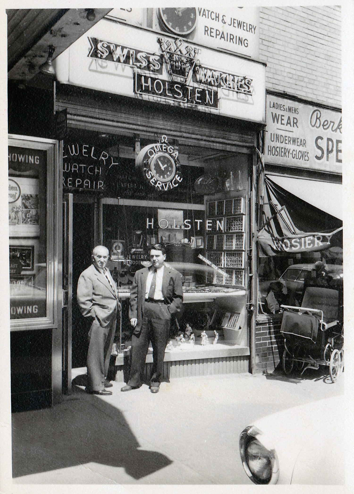 black and white photo of two men standing in front of a jewelry and watch repair shop that says "holsten" on the sign
