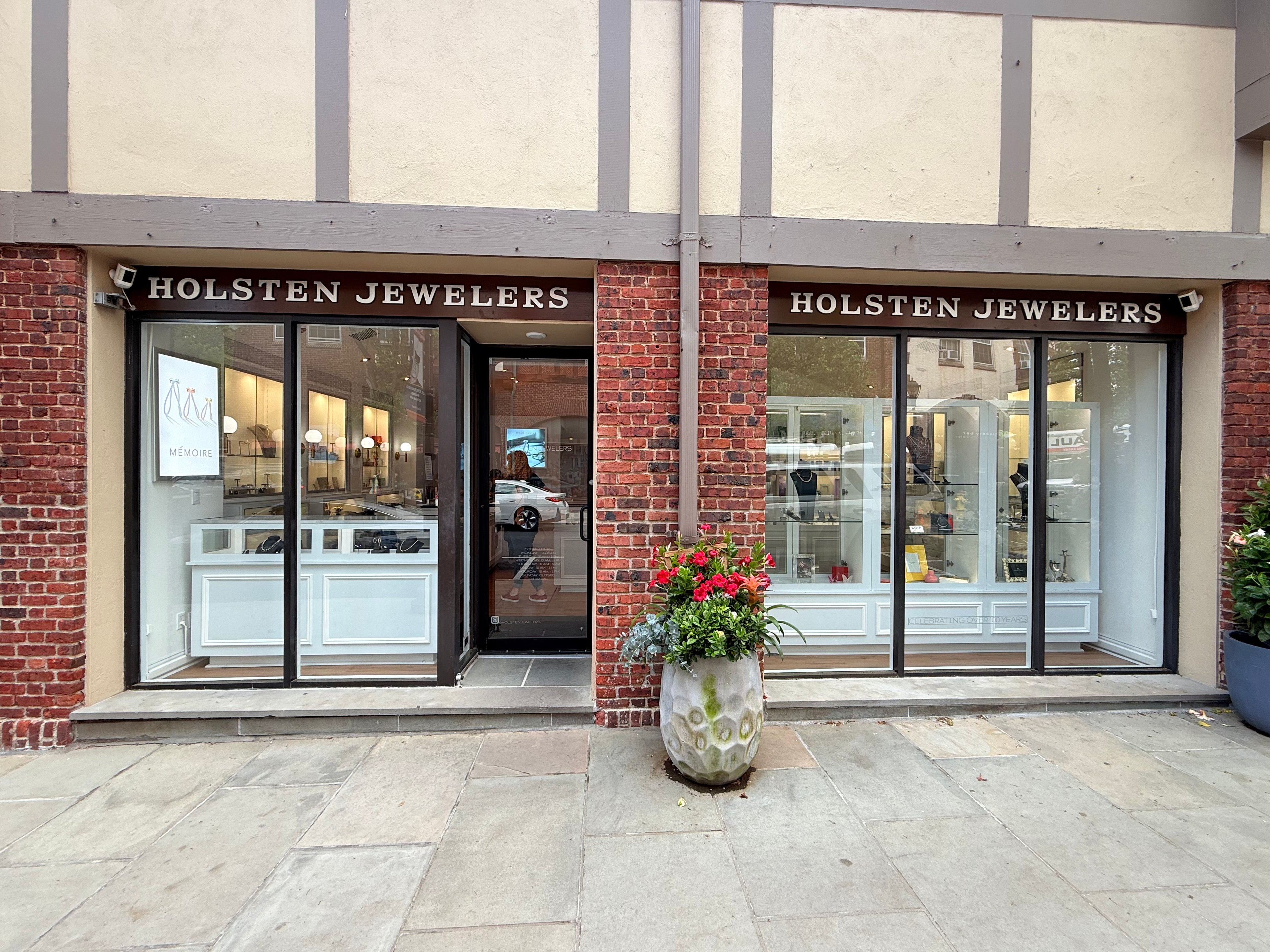 Brick storefront with dark brown window panes and a large flower pot with two signs that read "Holsten Jewelers"