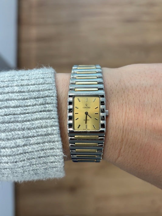 Silver square-shaped watch with a gold face on the wrist of someone wearing a grey sweater against a blurred wood background
