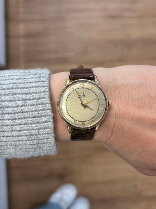 Gold watch with a black snakeskin leather band on the wrist of someone wearing a grey sweater against a blurred wood background