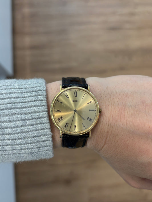Piaget men's watch with a yellow gold face and black band on the wrist of someone wearing a grey sweater against a blurred wood background